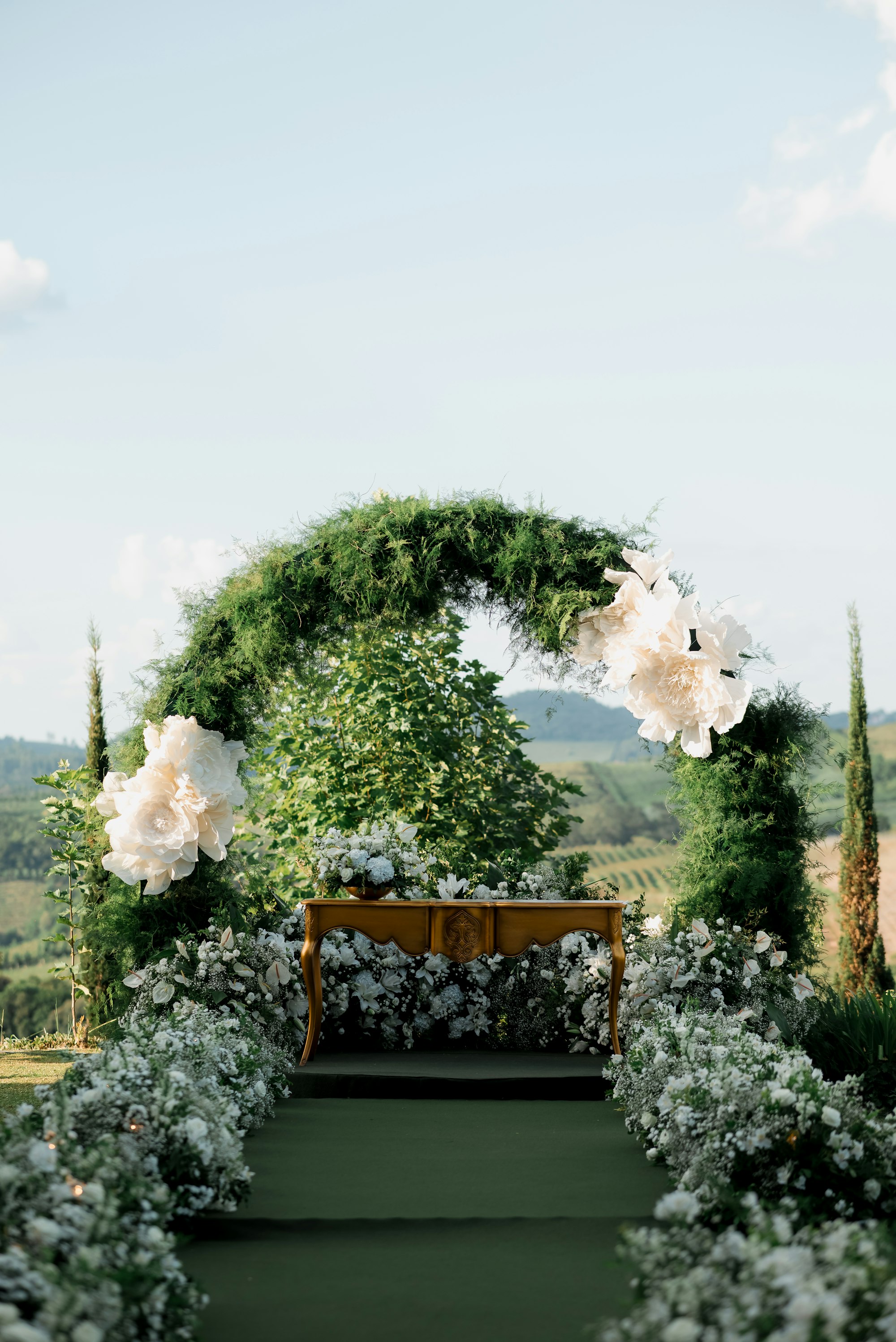 An outdoor wedding ceremony arch dressed in greenery and white florals, overlooking open countryside.